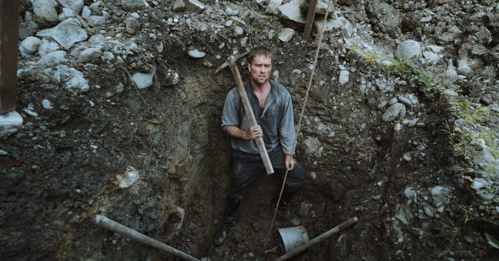 A rugged miner stands in a deep excavation holding a pickax, surrounded by rocky terrain.