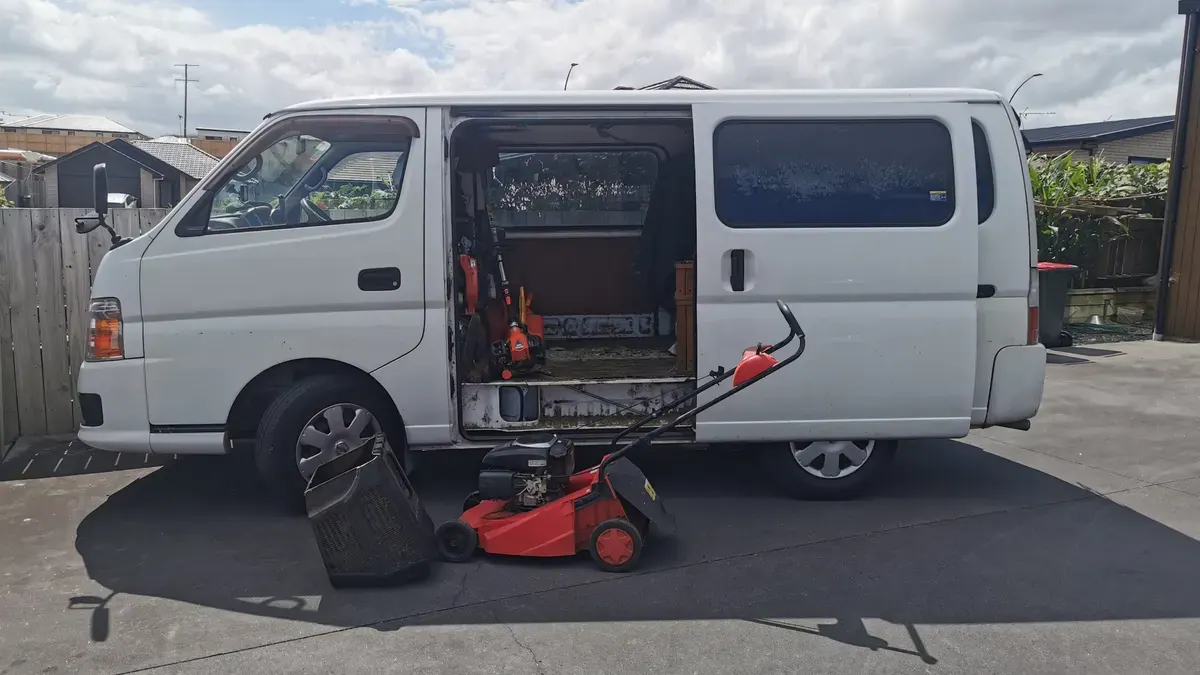 White work van parked on a street with the side door open, trimmers inside and a red mower and catcher on the road beside it