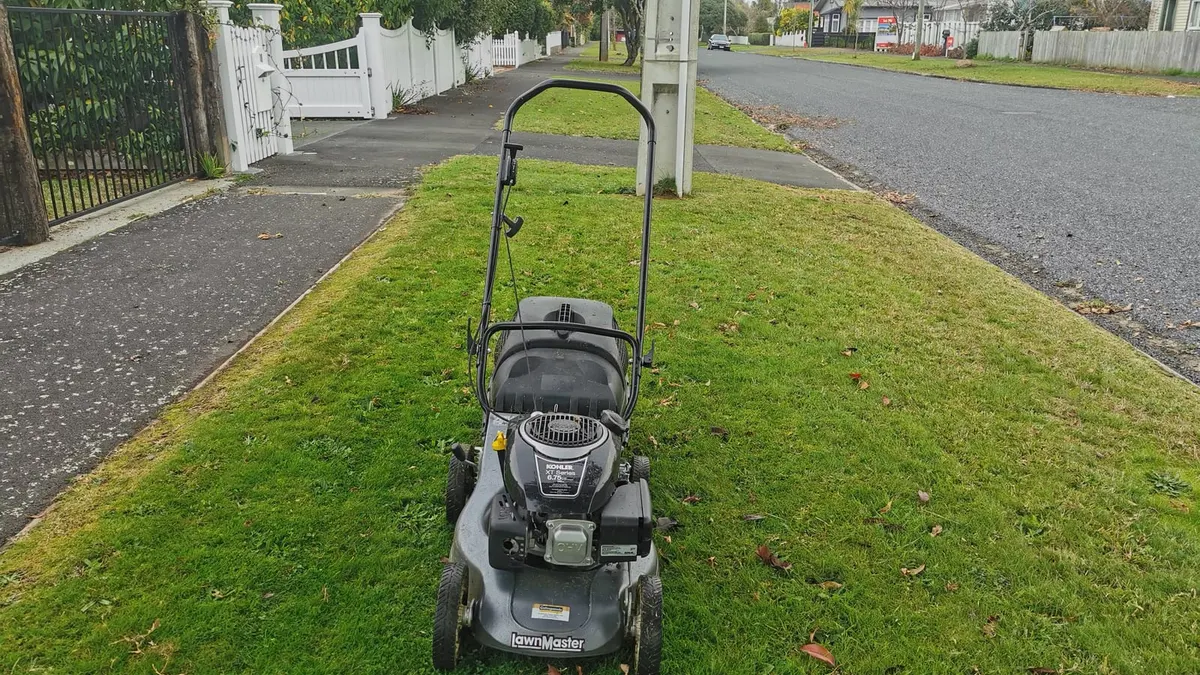 A push mower on a mown grass berm beside a footpath, with a long straight street of houses and front lawns behind it