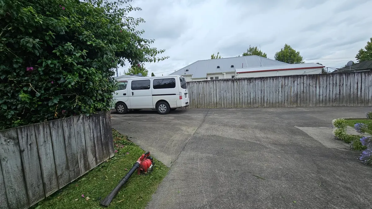 A white work van parked in a concrete driveway behind a timber fence, with a red leaf blower lying on a freshly edged strip of lawn